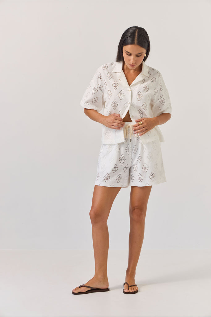 Woman wearing a white embroidered outfit on a plain background. Tuesday Label revere Top and Umpire Shorts in Diamond.