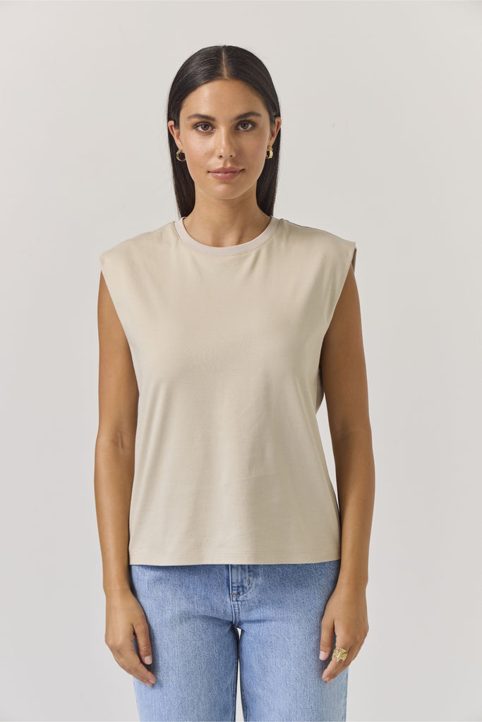 Woman wearing a beige sleeveless tank top and blue jeans on a plain background. Tuesday Noon Tank in Cement.