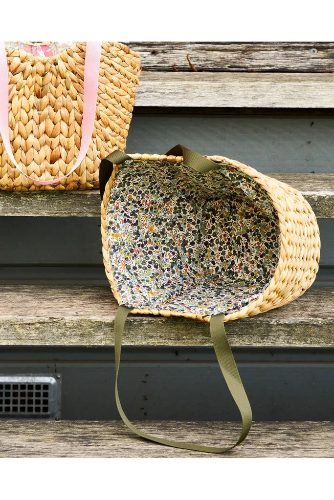 Robert Gordon Market Lane Wiltshire Basket: a water hyacinth woven basket with green canvas shoulder handles, lined with a green and blue floral Liberty print fabric. Basket shown laying on a wooden step on its side so enabling viewer to see the beautiful interior Liberty lining.