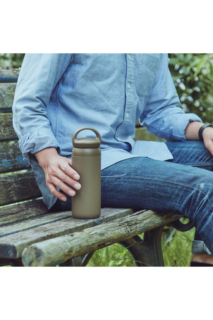 Kinto Day Off Khaki Insulated Tumbler. A person sitting on a wooden bench holding the  insulated bottle/tumbler outdoors.