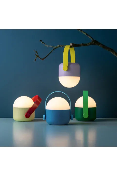 Remember OLE LED Portable Lamps in 4 colourful colourways. A Lilac and Yellow lamp hanging by the handle from a branch, and the three other colourways, Sand & Red, Blue & Blue and Green & Green, sit underneath it. 
