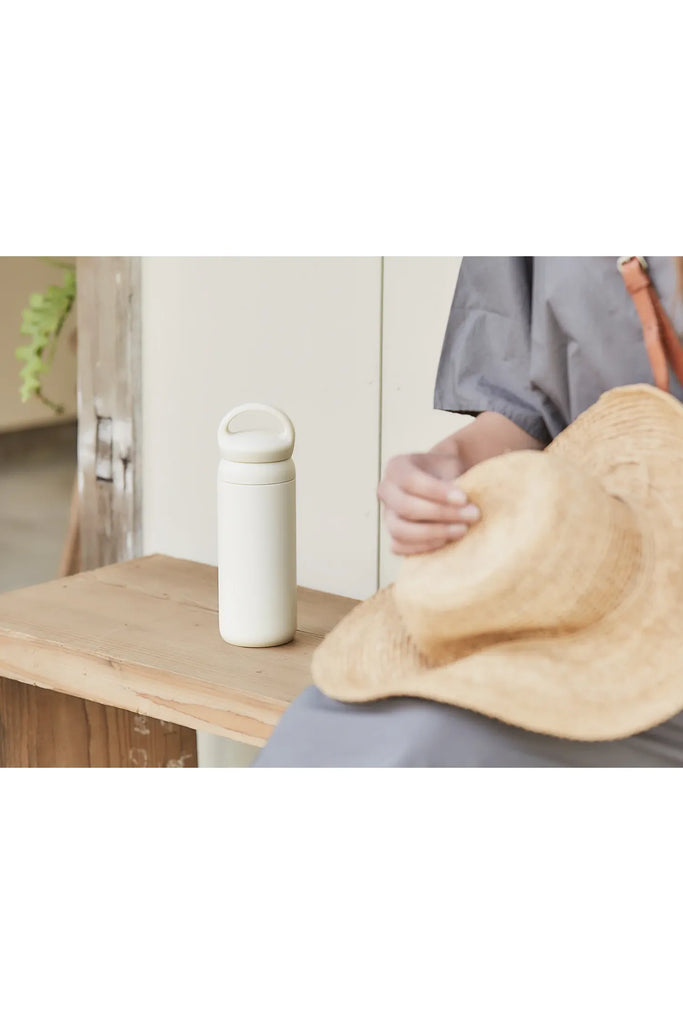 Kinto Day Off Tumbler in Sand Beige. Beige water bottle with a handle on a wooden surface with a person holding a straw hat in the foreground.