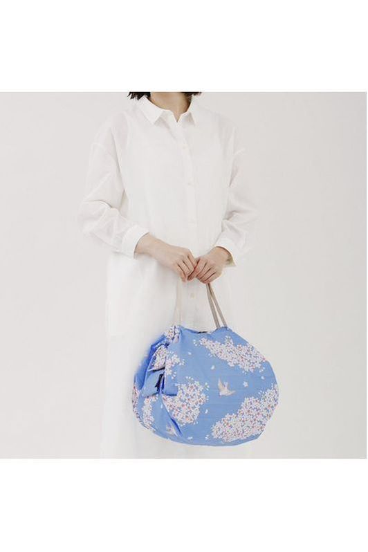 Shupatto Compact Reusable Bag Hana Ni Tore from the Japan Collection. Person holding the Hana Ni Tore reusable compact medium-size blue floral-patterned bag against a white background.