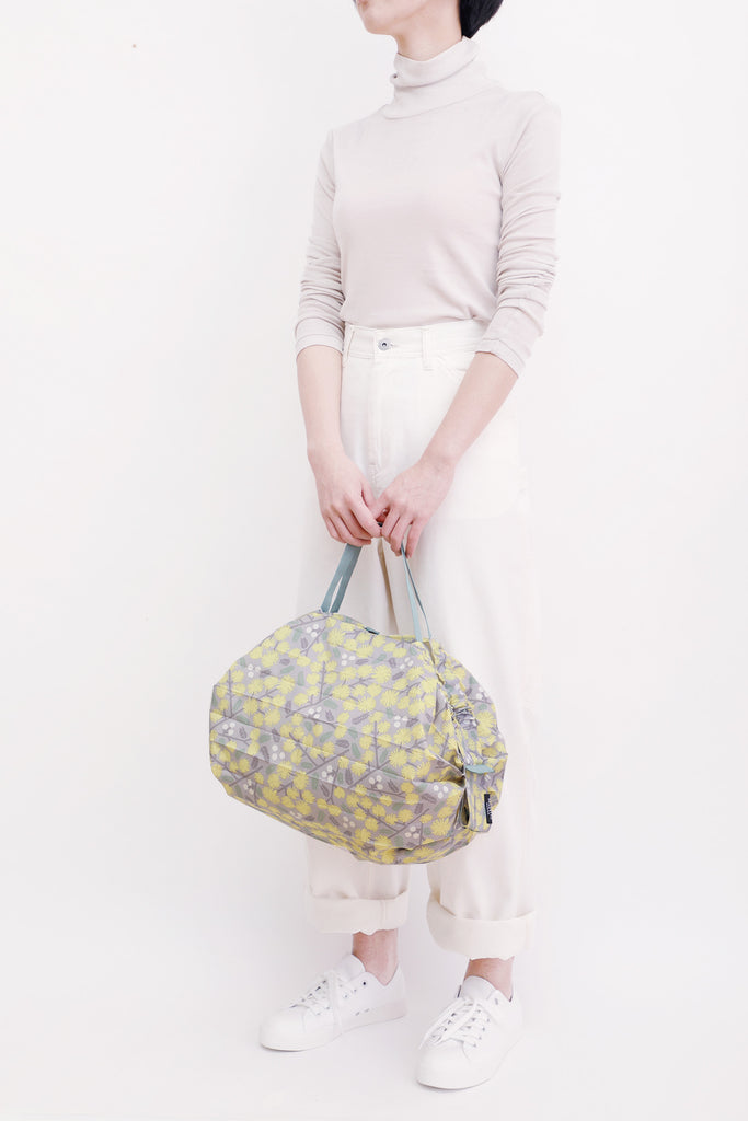 Shupatto Compact Medium reusable shopping bag featuring a floral pattern in grey, yellow, and sage green on a grey background, complemented by a sage green strap. A person is seen holding the floral-patterned bag against a white background.
