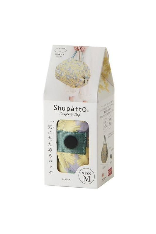 The Shupatto Compact Medium reusable shopping bag in the "Hana" design features a floral pattern in grey, yellow, and sage green on a grey background, complemented by a sage green strap. The image shows the bag in s display box with a cut out window so the bag pattern is able to be viewed.