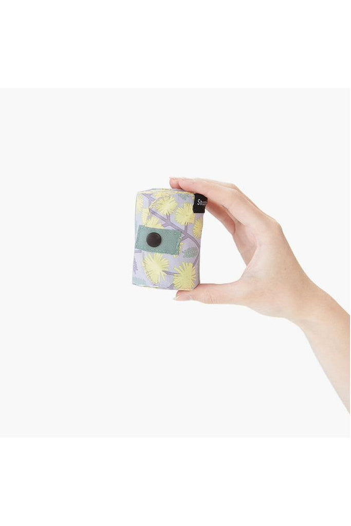 The Shupatto Compact Medium reusable shopping bag, called Hana, features a floral pattern in grey, yellow, and sage green against a grey background, complemented by a sage green strap. The bag can be rolled up into a small cylindrical shape and secured with a dome for easy storage.