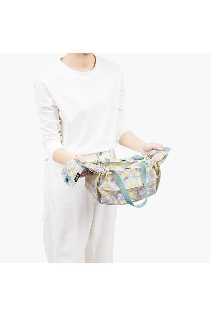 The Shupatto Compact Medium reusable shopping bag in the "Hana" design features a floral pattern in grey, yellow, and sage green on a grey background, complemented by a sage green strap. In the image, a person is holding the bag and pulling both ends to elongate it, preparing to roll it into a cylindrical shape for convenient storage.