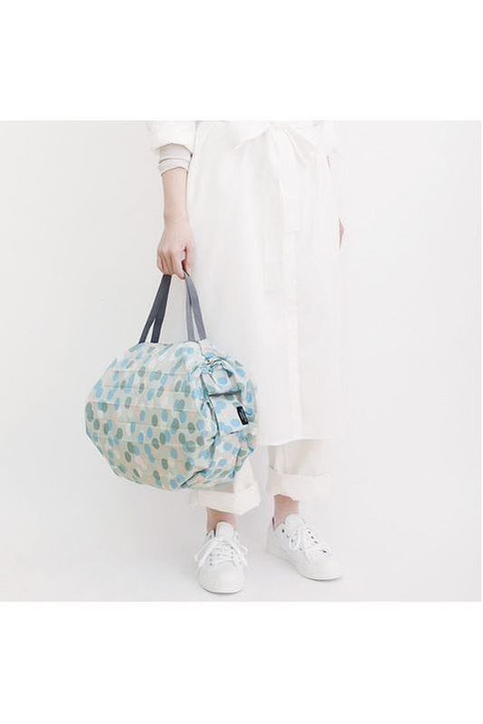 A person is holding a Shupatto Compact medium reusable shopping bag, Arare, featuring a hailstone pattern in light blue, sage green, cream, and grey handles against a plain background.