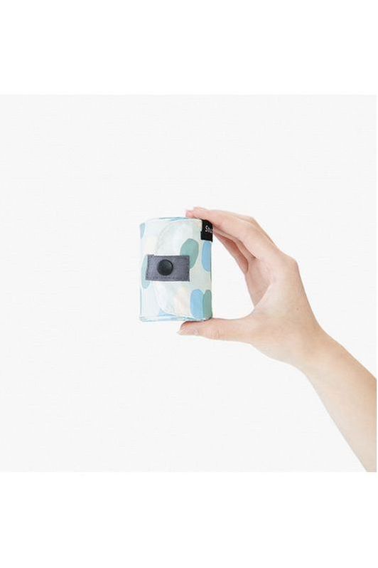 Shupatto Compact medium reusable shopping bag, Arare, featuring a hailstone pattern in light blue, sage green, cream, and grey handles. A hand holds the bag rolled up into a small cylindrical shape against a white background.