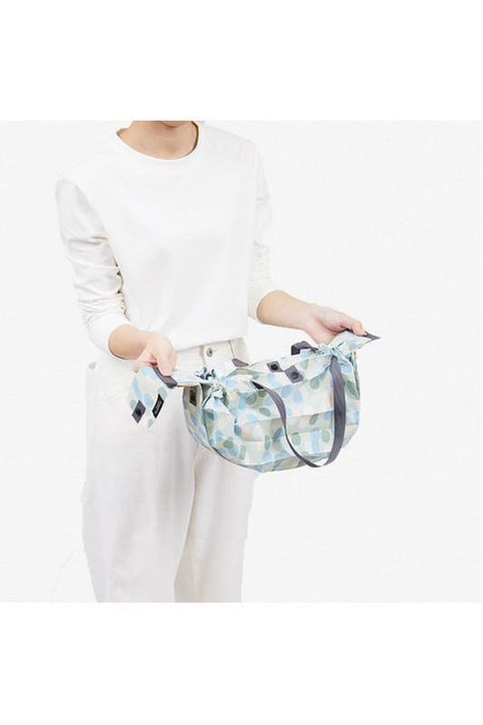 Shupatto Compact medium reusable shopping bag, Arare, features a hailstone pattern in light blue, sage green, cream, and grey handles. A person holds the patterned bag, pulling it straight out to roll it into a small cylindrical shape.