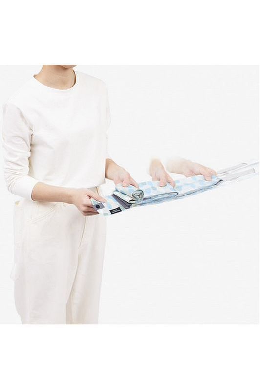 The Shupatto Compact Medium Reusable Shopping Bag in Arare features a hailstone pattern in light blue, sage green, cream, and grey handles. A person is shown holding the bag, which can be compressed and then pulled straight and is being rolled into a small cylindrical shape for easy storage. 